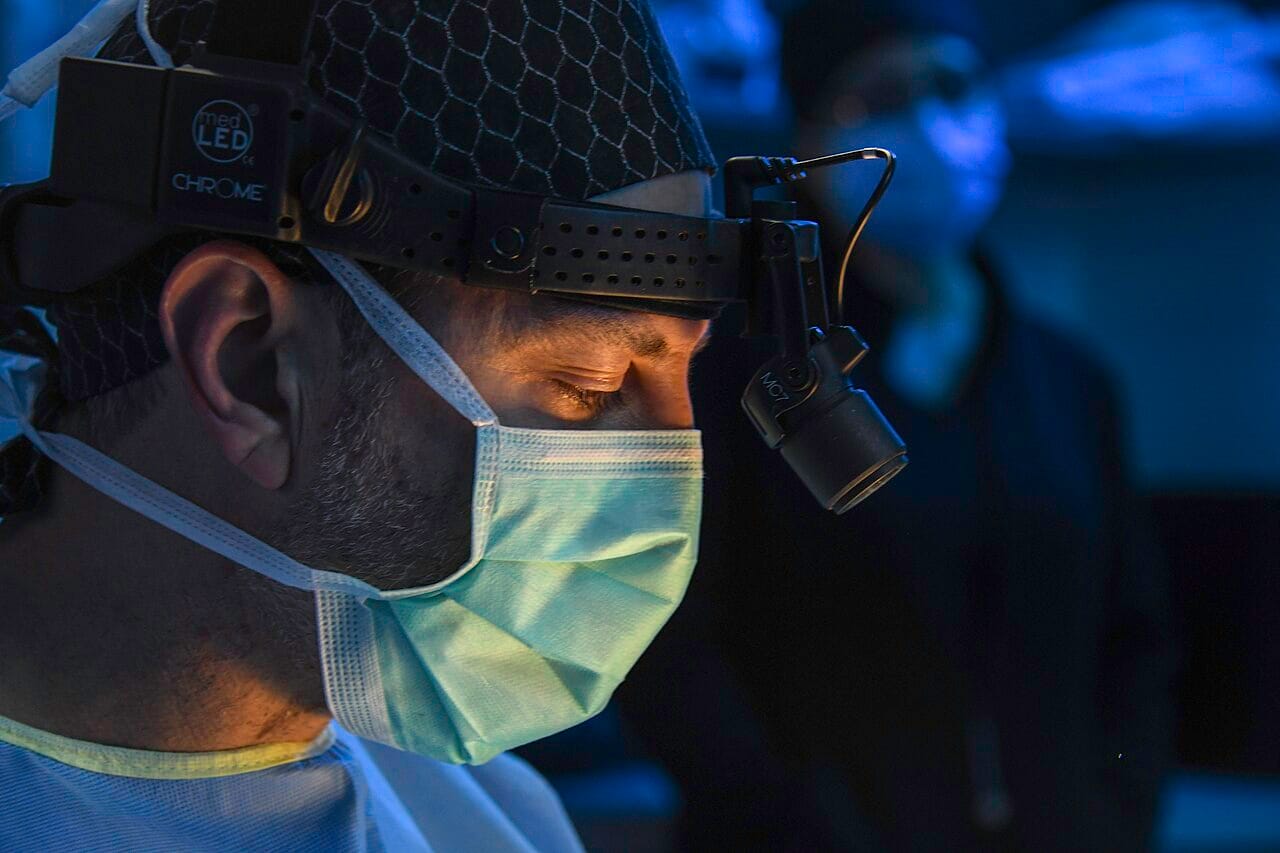 A surgeon in an operating room, in dark blue-green scrubs under theatre lighting.