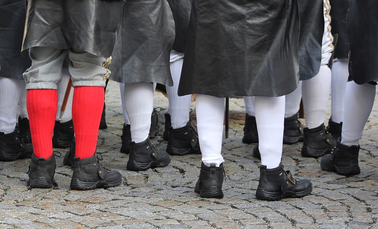 A row of legs at a parade: white stockings, red stockings, black shoes.