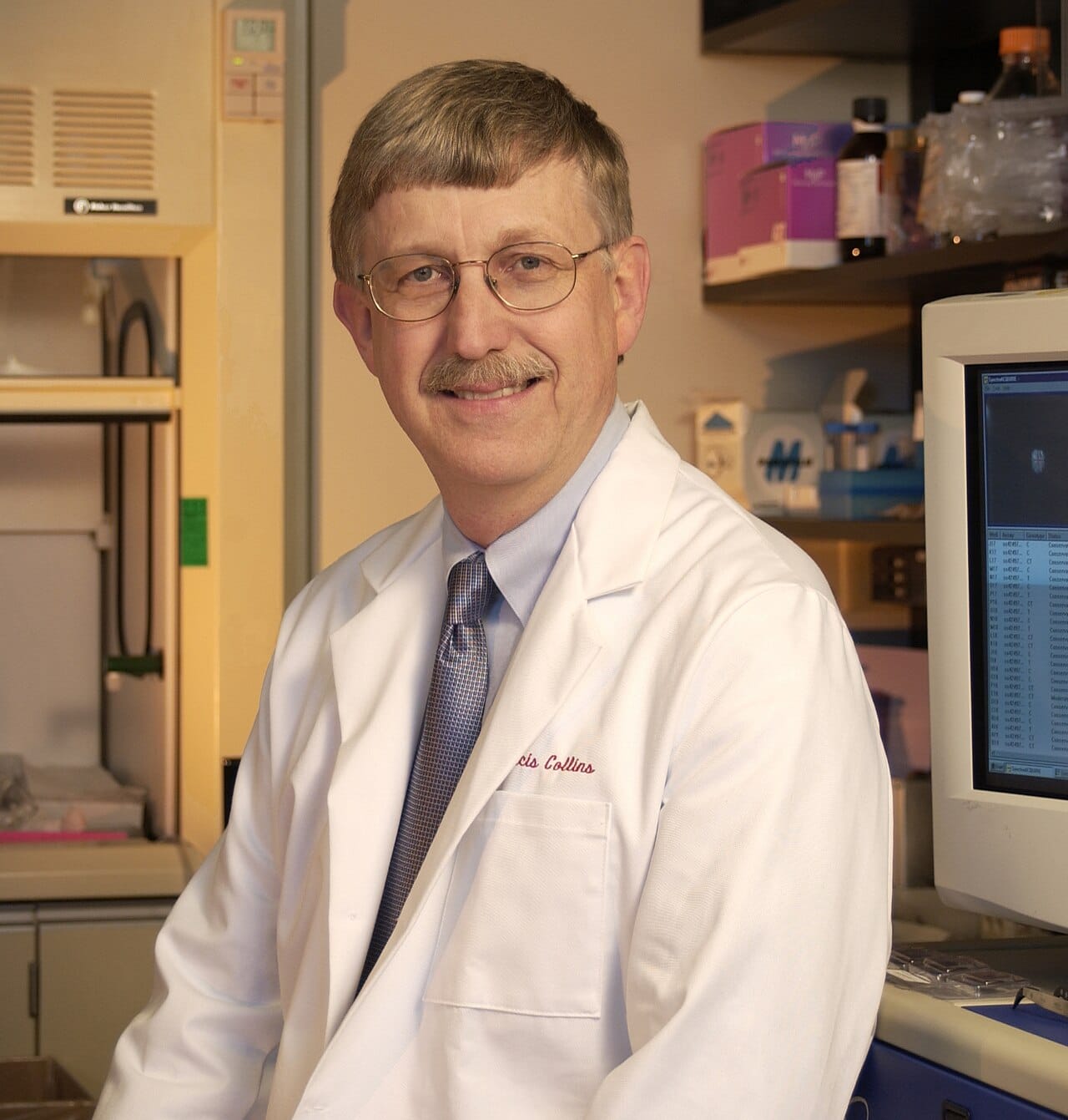A director photographed in a white coat over a shirt and tie.