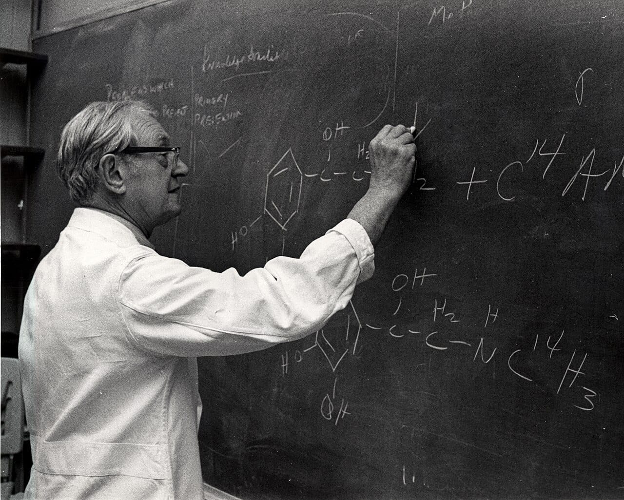 A biochemist at a blackboard, 1973, coat open over ordinary clothes.